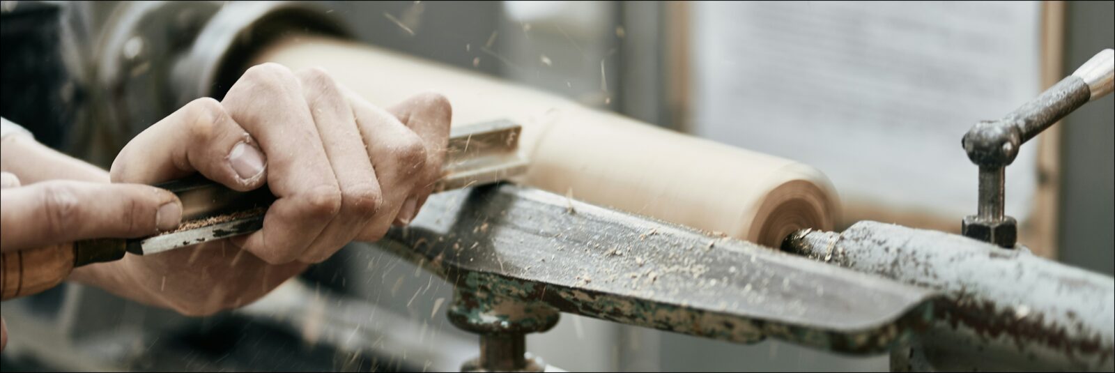 Close view of hands turning a wooden spindle on a lathe, chisel resting on the tool rest, shavings in the air